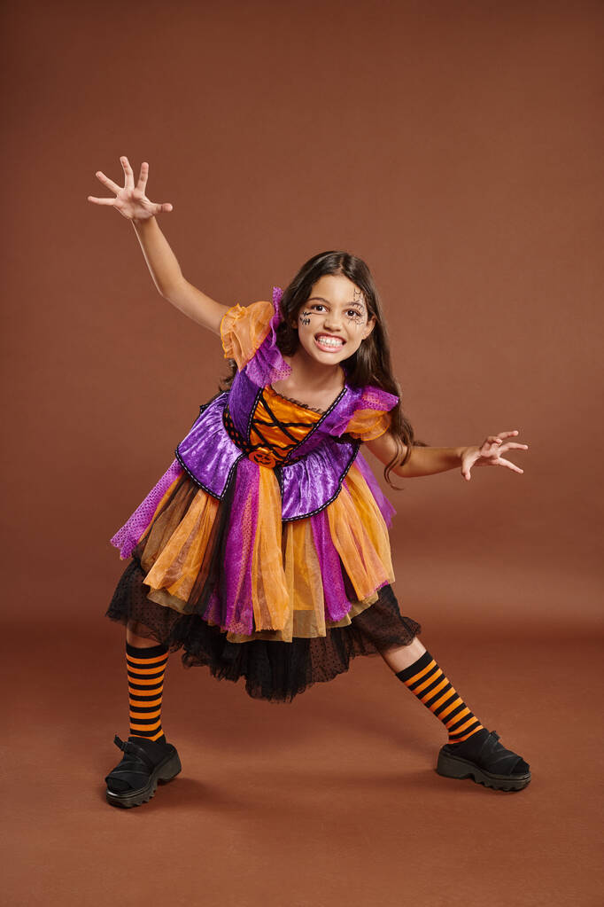 chica espeluznante en disfraz de bruja de Halloween gruñendo y haciendo gestos en el fondo marrón, longitud completa - Foto, Imagen
