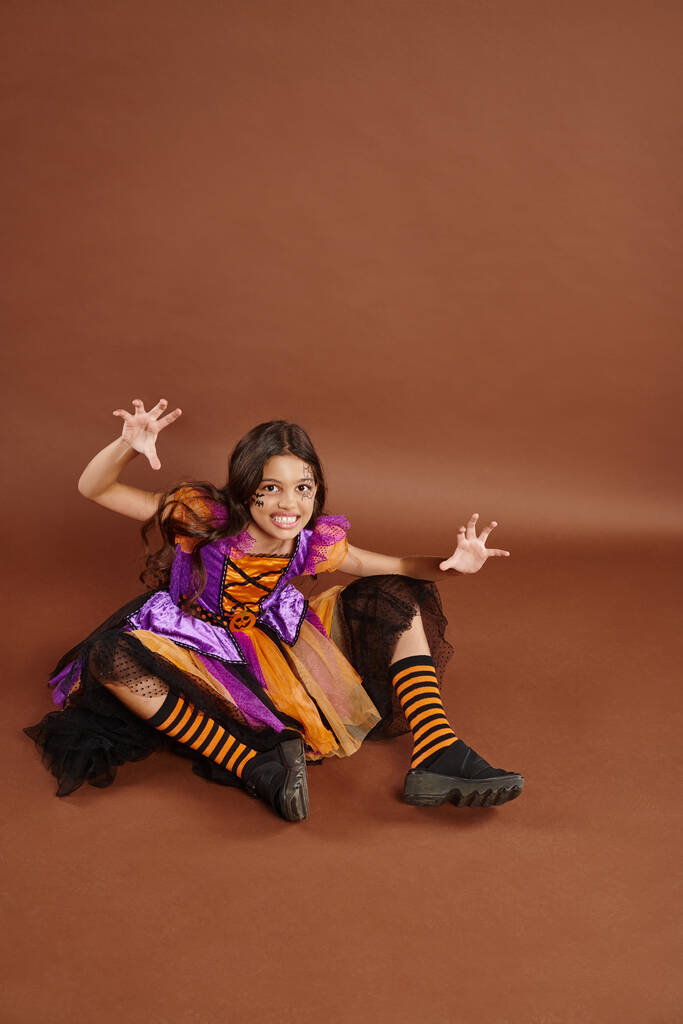 kísérteties lány Halloween boszorkány jelmez ül és morgó barna háttér, október 31 - Fotó, kép