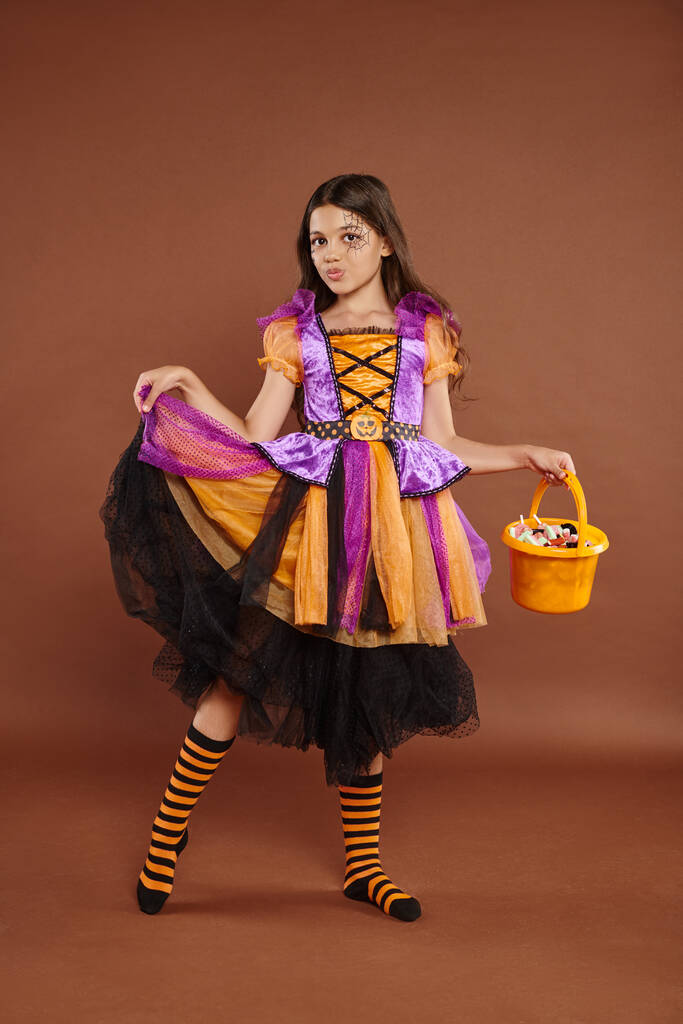 rozkošná dívka v halloweenském kostýmu drží kbelík s bonbóny a drží sukni na hnědém pozadí - Fotografie, Obrázek