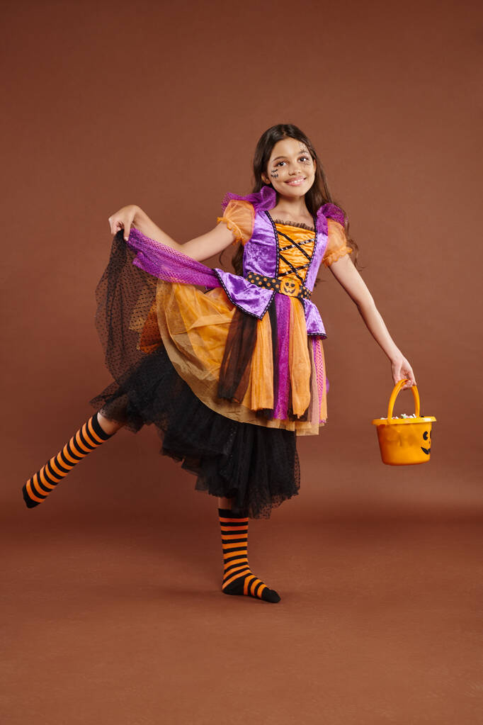 fröhliches Mädchen im Halloween-Kostüm hält Eimer mit Bonbons und hält Rock auf braunem Hintergrund - Foto, Bild