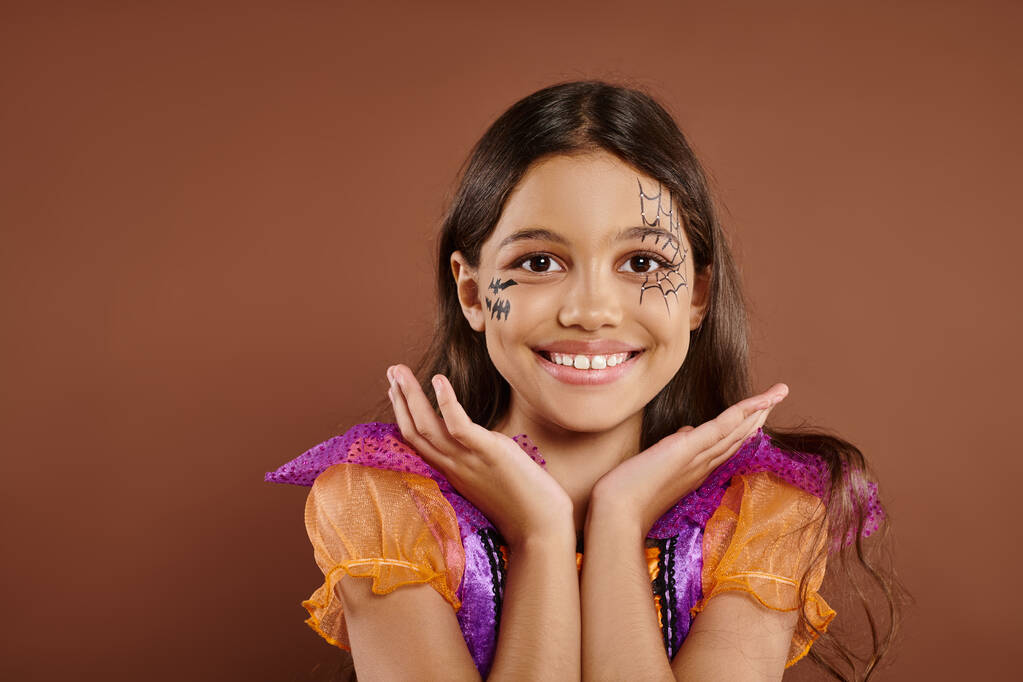 vrolijk meisje in Halloween kostuum en spinnenweb make-up gebaar op bruine achtergrond, truc of traktatie - Foto, afbeelding