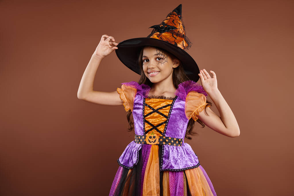 happy girl in Halloween costume and pointed hat posing on brown background, little witch attire - Photo, Image