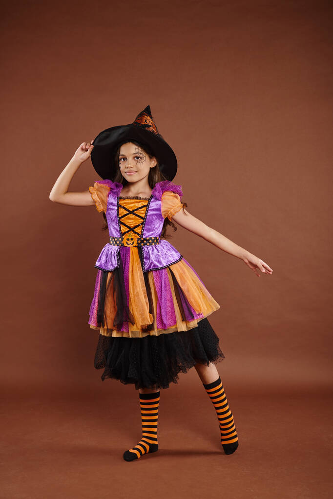 elegant meisje in Halloween kostuum en puntige hoed poseren op bruine achtergrond, gelukkig kleine heks - Foto, afbeelding