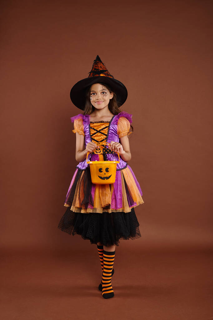 boldog lány halloween jelmez és hegyes kalap áll cukorka vödör barna háttér - Fotó, kép