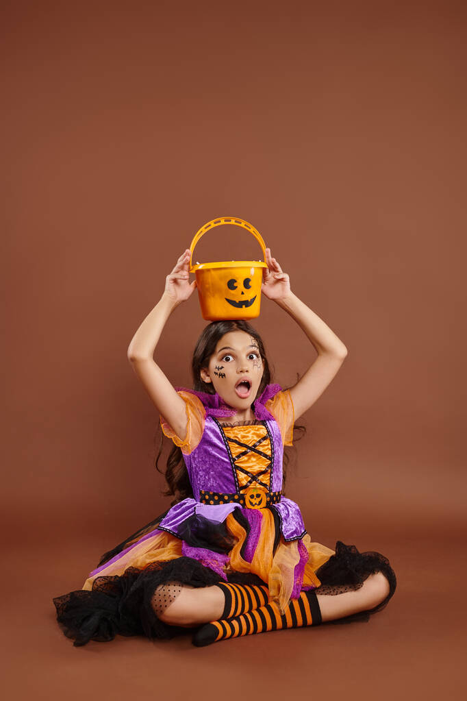 astonished girl in Halloween costume holding candy bucket on head and sitting on brown background - Photo, Image