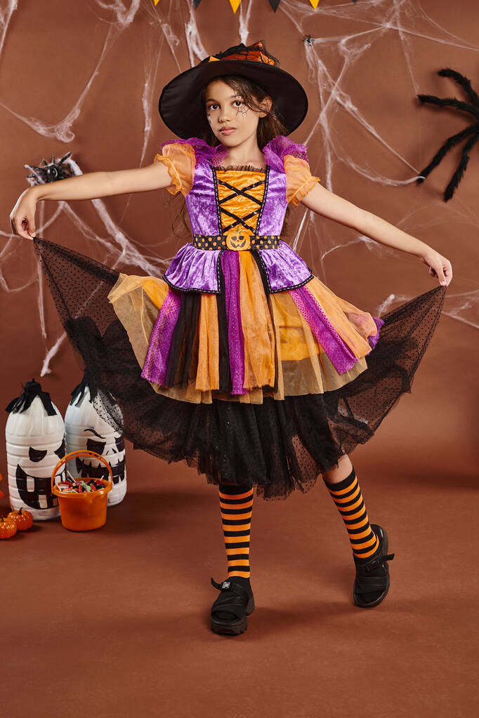 chica en sombrero de bruja y vestido sosteniendo falda cerca de cubo con dulces en marrón, concepto de Halloween - Foto, imagen