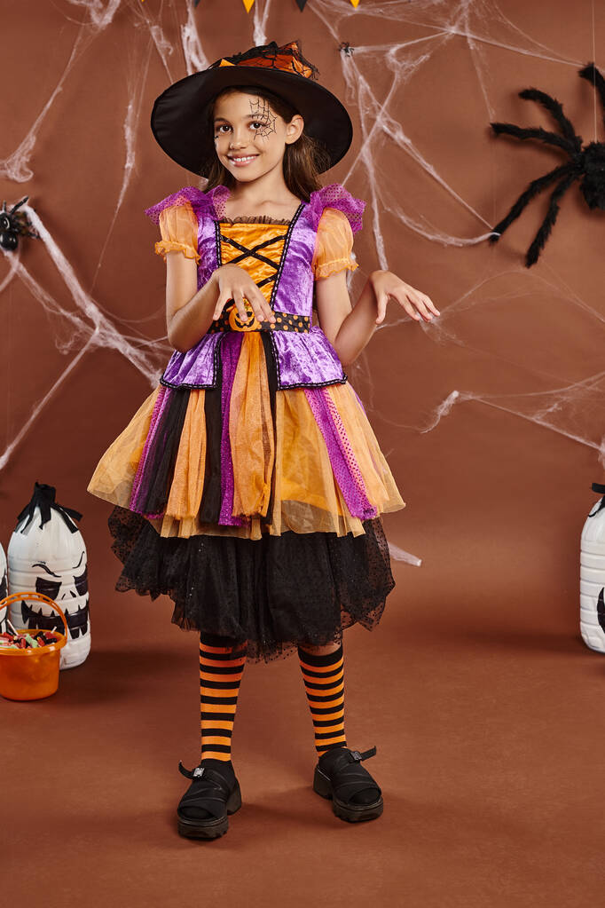 wesoła dziewczyna w kapeluszu czarownicy i sukienka taniec w pobliżu wiadra ze słodyczami na brązowym, koncepcja Halloween - Zdjęcie, obraz