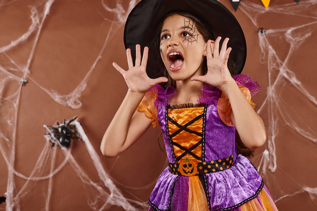 chica emocional en sombrero de bruja y disfraz de Halloween gritando y haciendo gestos sobre fondo marrón - Foto, imagen