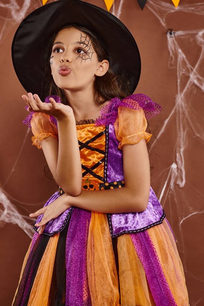 cute girl in witch costume and pointed hat sending air kiss on brown backdrop, Halloween concept - Photo, Image