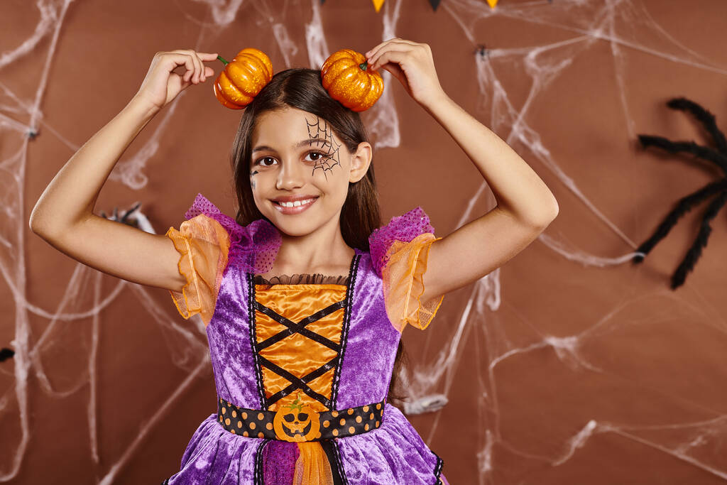 chica divertida con maquillaje de tela de araña sonriendo y sosteniendo calabazas cerca de la cabeza en el fondo marrón, Halloween - Foto, Imagen