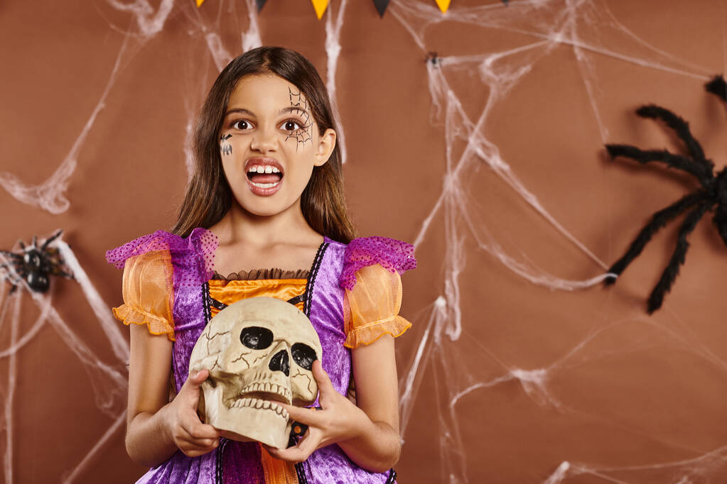 fille en costume d'Halloween tenant le crâne et grognant à la caméra sur fond brun, saison effrayante - Photo, image
