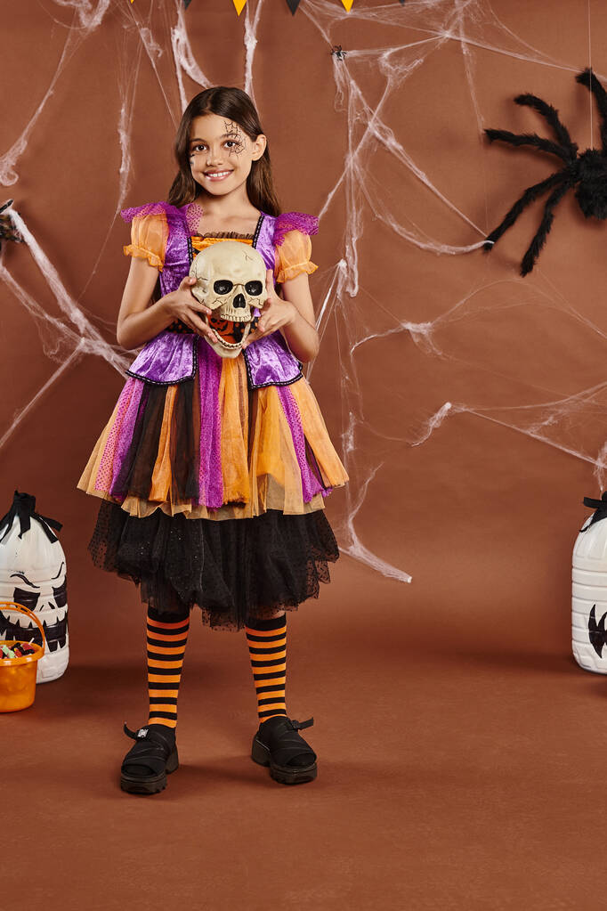 cheerful girl in dress holding skull and smiling on brown background, Halloween spooky season - Photo, Image