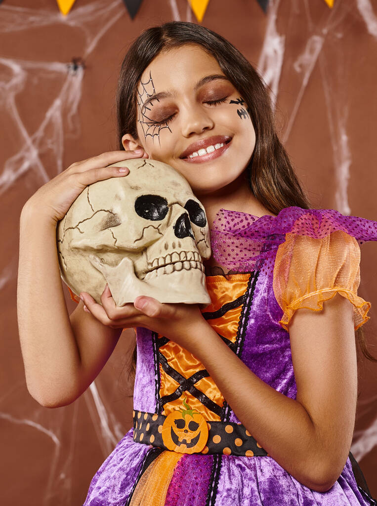chica positiva en vestido sosteniendo cráneo y sonriendo sobre fondo marrón, Halloween temporada espeluznante - Foto, Imagen