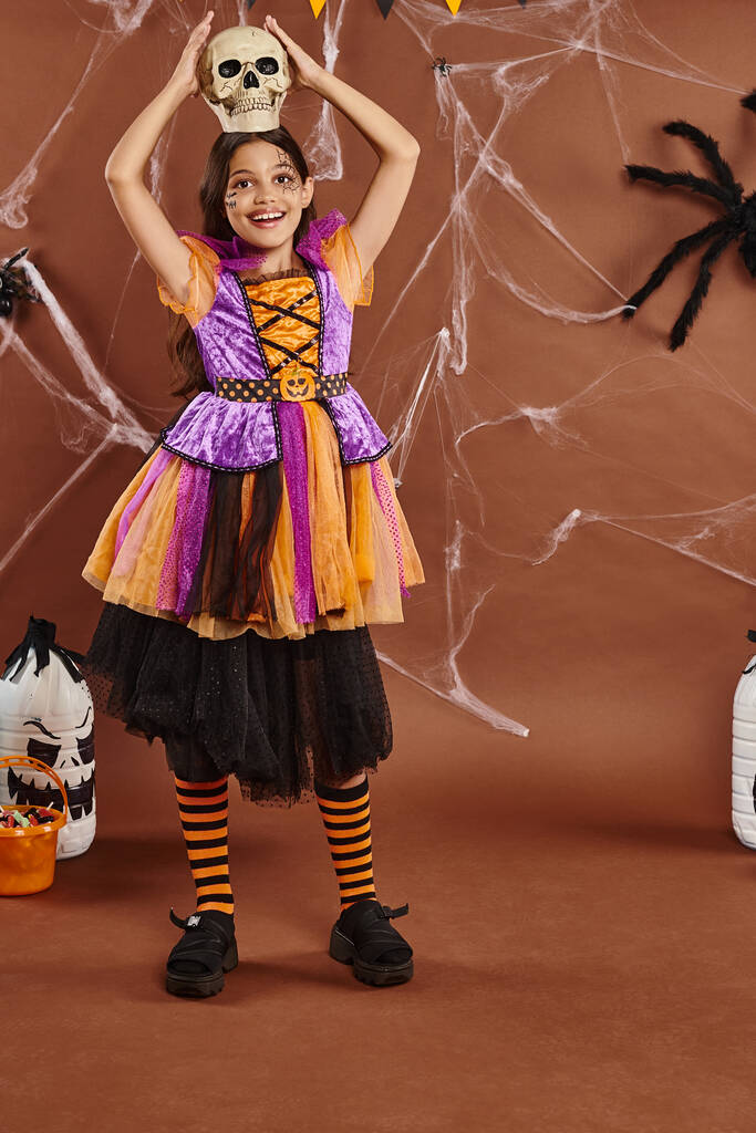 joyful girl in Halloween dress standing with skull on head on brown background, spooky season - Photo, Image