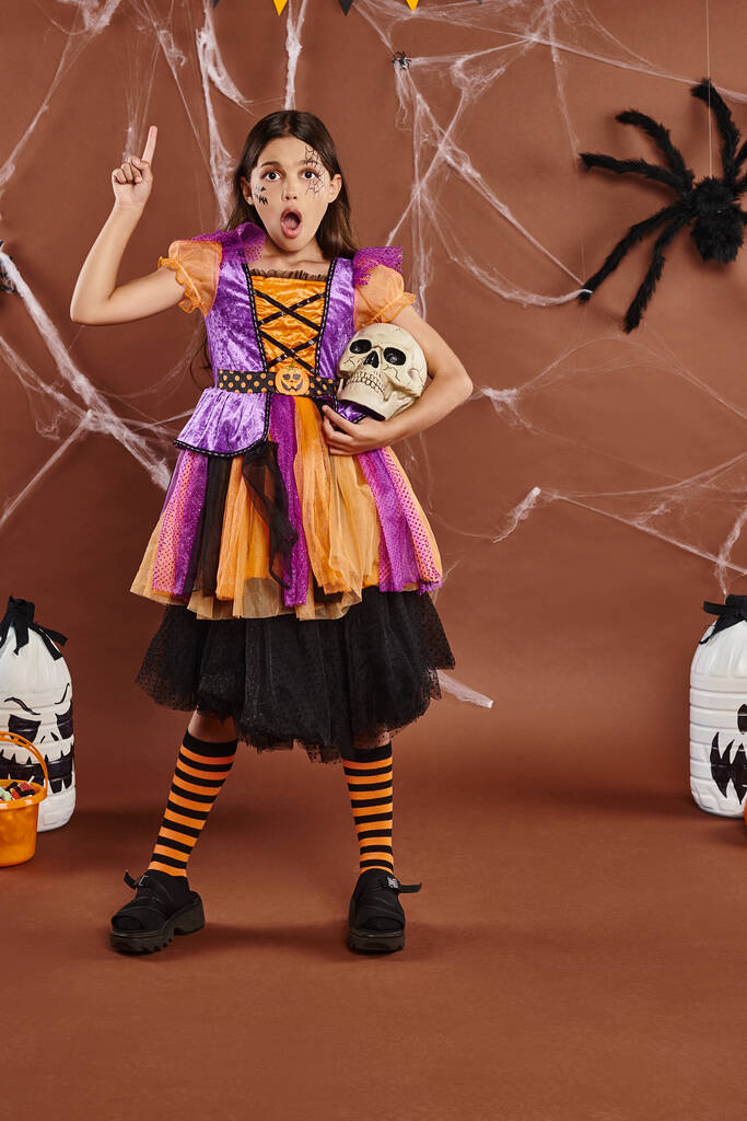 σοκαρισμένη κοπέλα στο Halloween φόρεμα στέκεται με το κρανίο και δείχνει προς τα πάνω με το δάχτυλο σε καφέ φόντο - Φωτογραφία, εικόνα