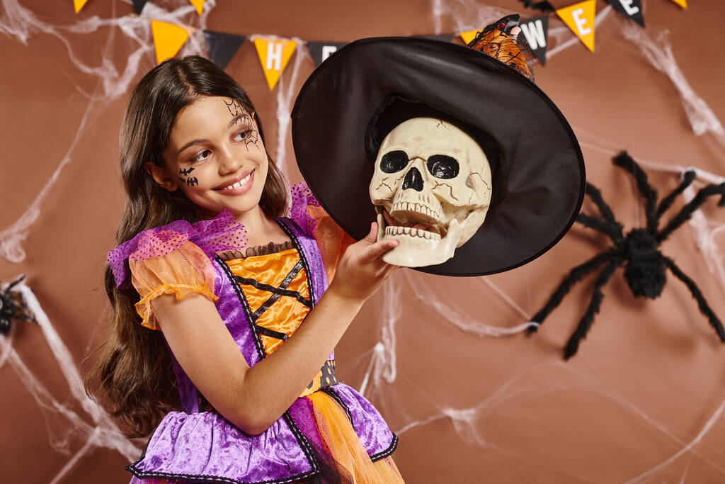 happy girl in Halloween attire wearing witch hat on skull on brown background, spooky season - Photo, Image