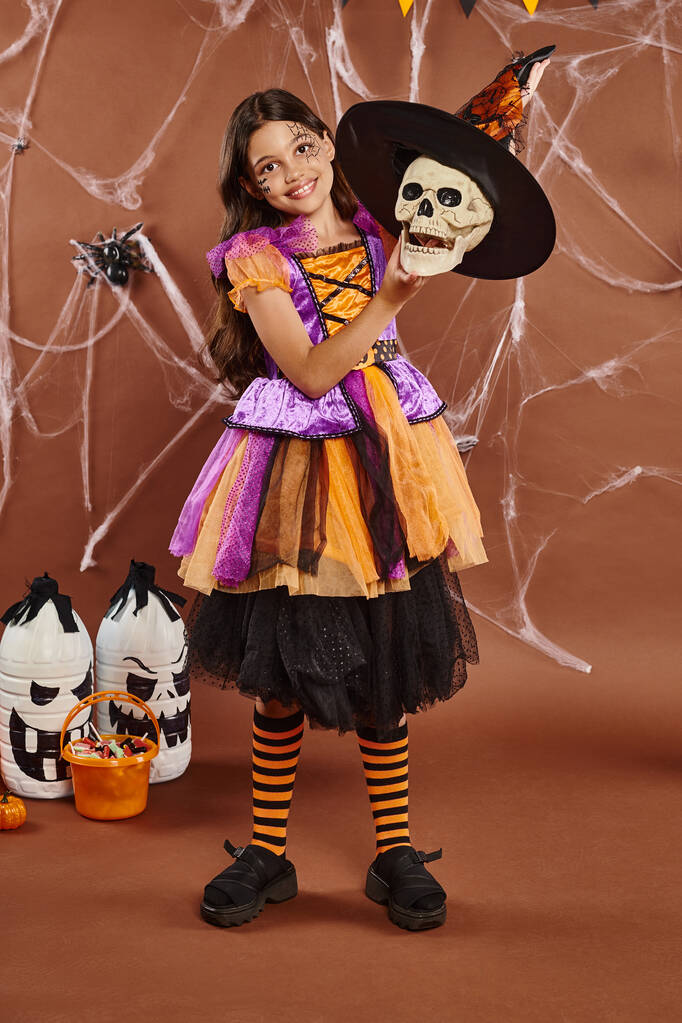 fröhliches Mädchen im Halloween-Kostüm mit Hexenhut auf Totenkopf auf braunem Hintergrund, gruselige Jahreszeit - Foto, Bild