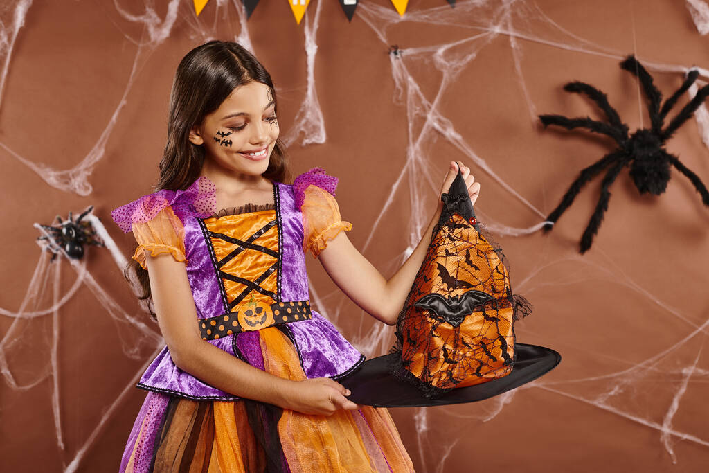 šťastná dívka v halloweenské čarodějnice kostým držení špičatý klobouk na hnědém pozadí, strašidelné období - Fotografie, Obrázek