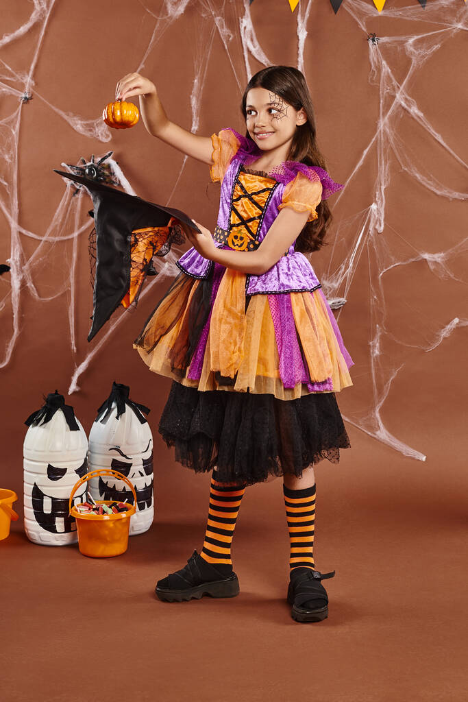 happy child in Halloween witch costume holding pumpkin near pointed hat on brown background - Photo, Image