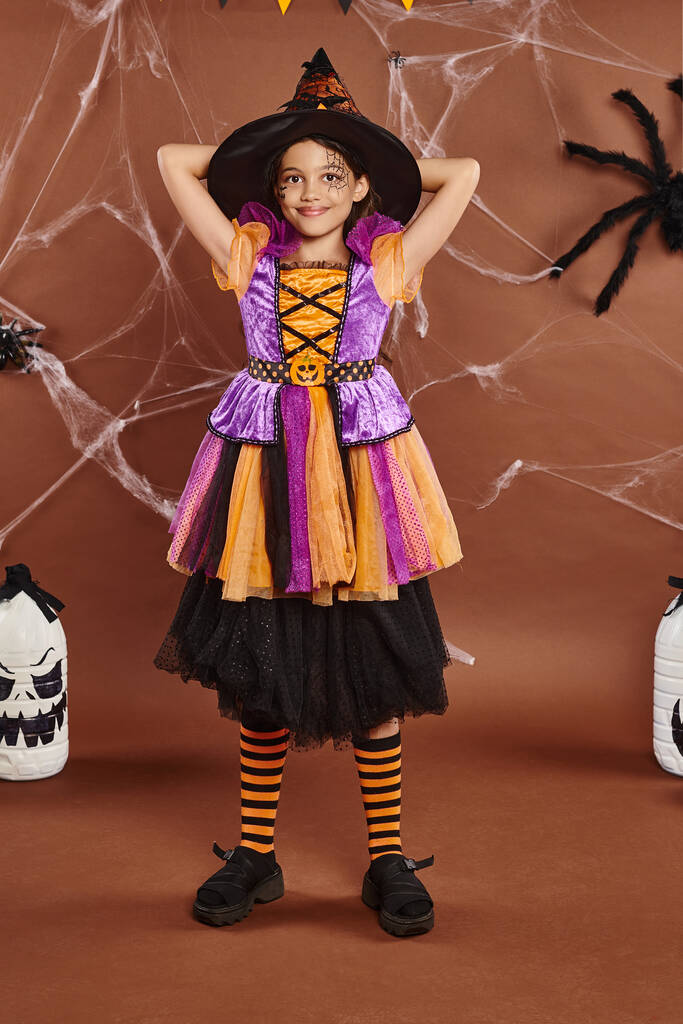 щаслива дівчина в капелюсі відьми і костюм Хеллоуїна, що стоїть поруч з солоденьким декором на коричневому фоні - Фото, зображення