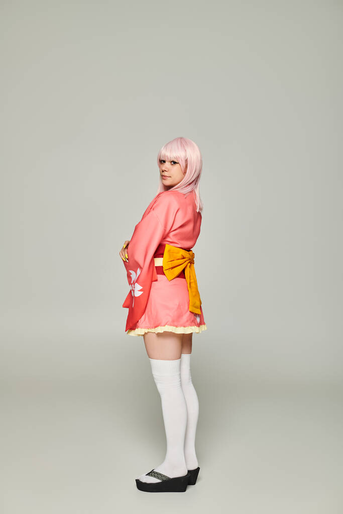 joven mujer de estilo anime en peluca rubia y kimono rosa con lazo amarillo sobre gris, concepto cosplay - Foto, Imagen