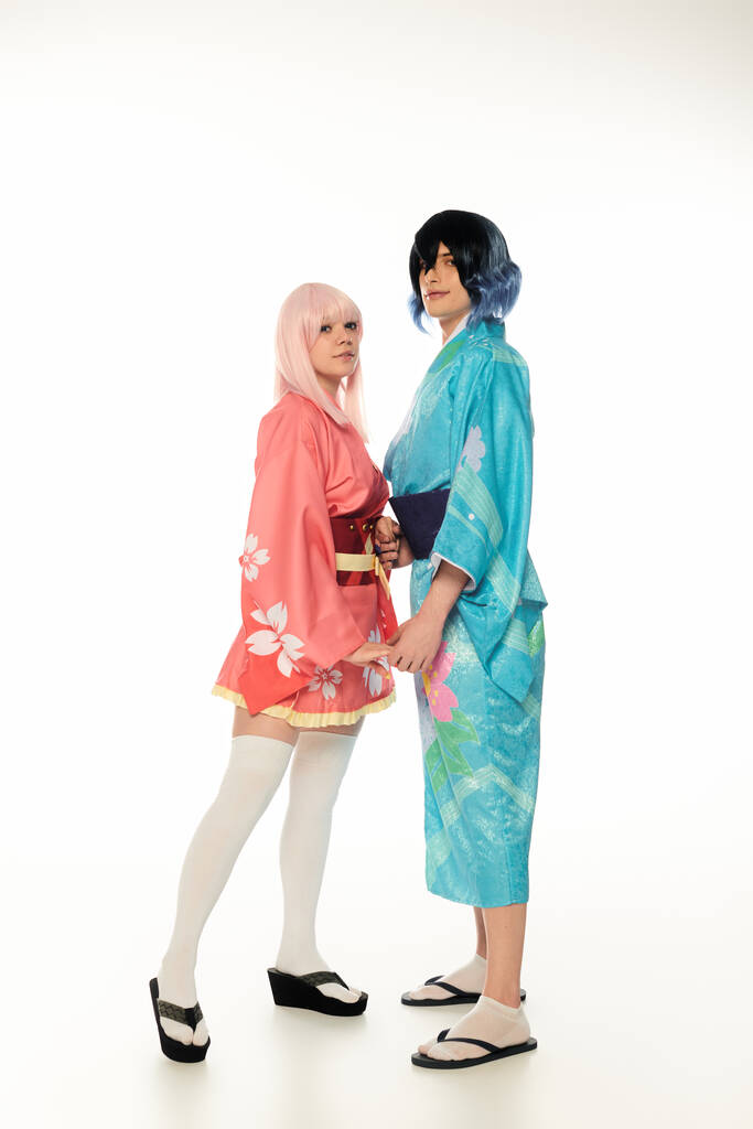 full length of young cosplayers in colorful kimonos holding hands on and looking at camera on white - Photo, Image