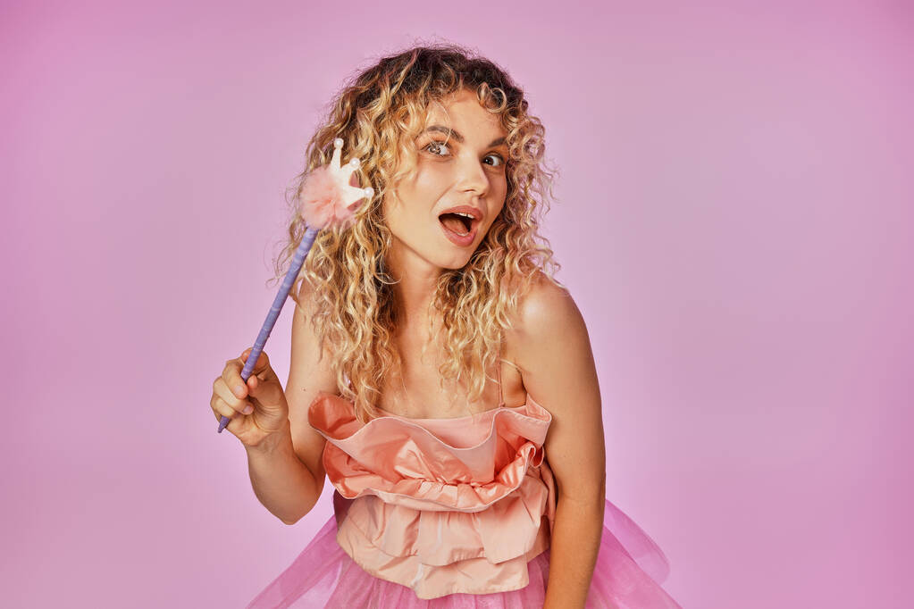 mujer rubia de pelo rizado en traje rosa de hada de los dientes sosteniendo varita mágica y mirando a la cámara - Foto, imagen