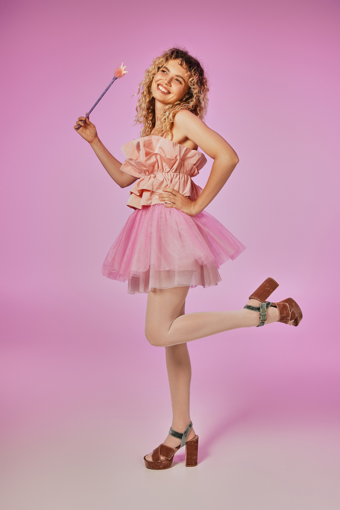 fröhliche blonde Frau mit lockigem Haar im Zahnfee-Kostüm, die auf einem Bein auf rosa Hintergrund steht - Foto, Bild