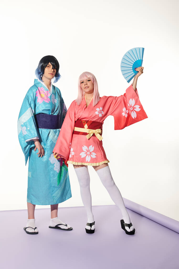 Anime stili kadın, kimonolu adamın yanında el fanı ile poz veriyor ve beyaz stüdyoda mor halıda peruk takıyor. - Fotoğraf, Görsel