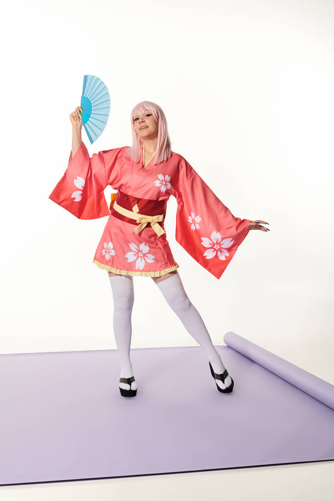 művészi cosplay nő rózsaszín kimono és szőke paróka kéz legyező lila szőnyeg fehér stúdió - Fotó, kép