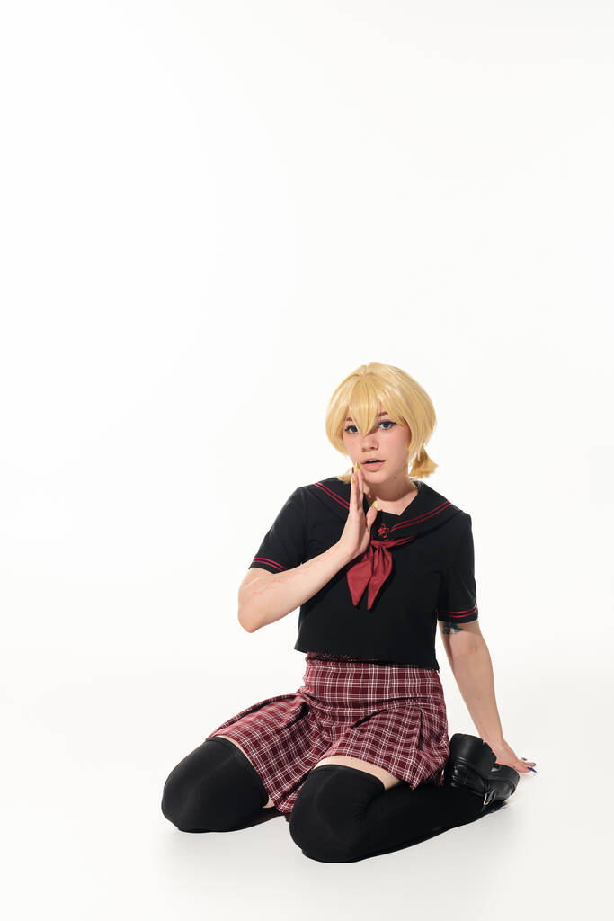 yellow blonde woman in trendy school unform sitting with hand near face on white, cosplay style - Photo, Image