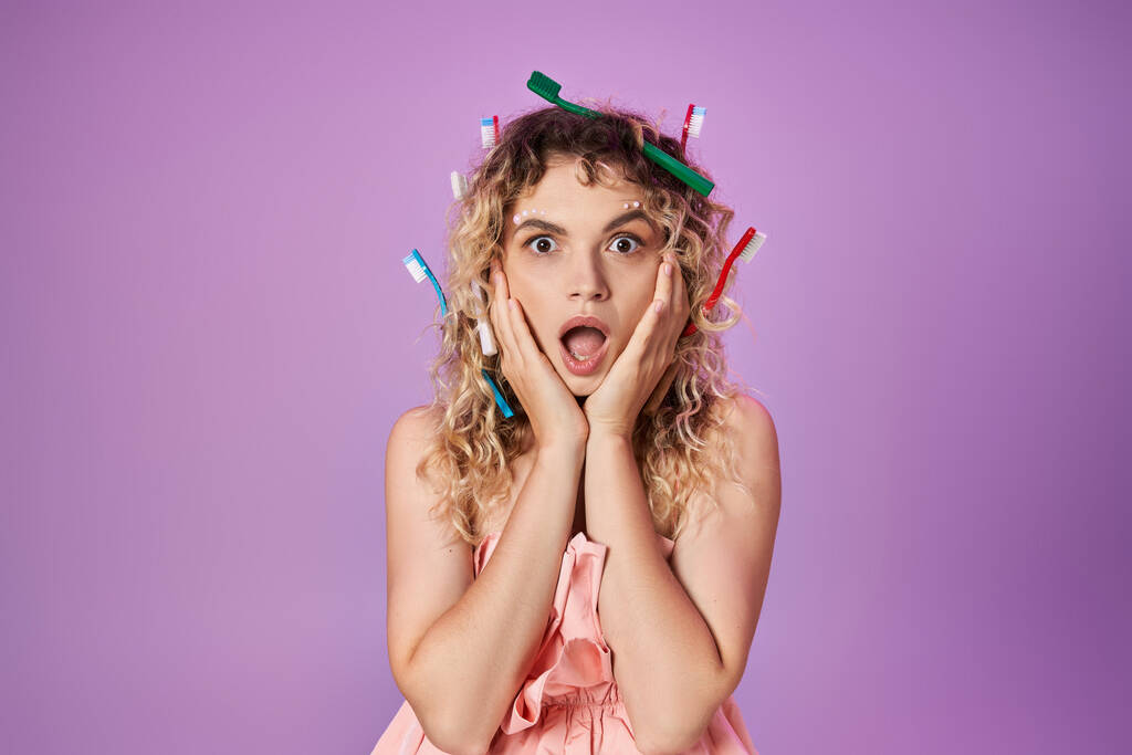 Schockierte Frau in rosa Kleid und Zahnbürsten im Haar, die Hände zu Wangen erhoben, Zahnfee-Konzept - Foto, Bild