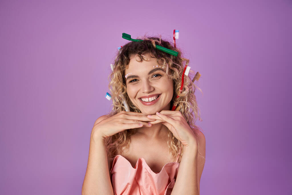 бодрая зубная фея в розовом наряде и зубных щетках в кудрявых волосах позирует с руками под подбородком - Фото, изображение