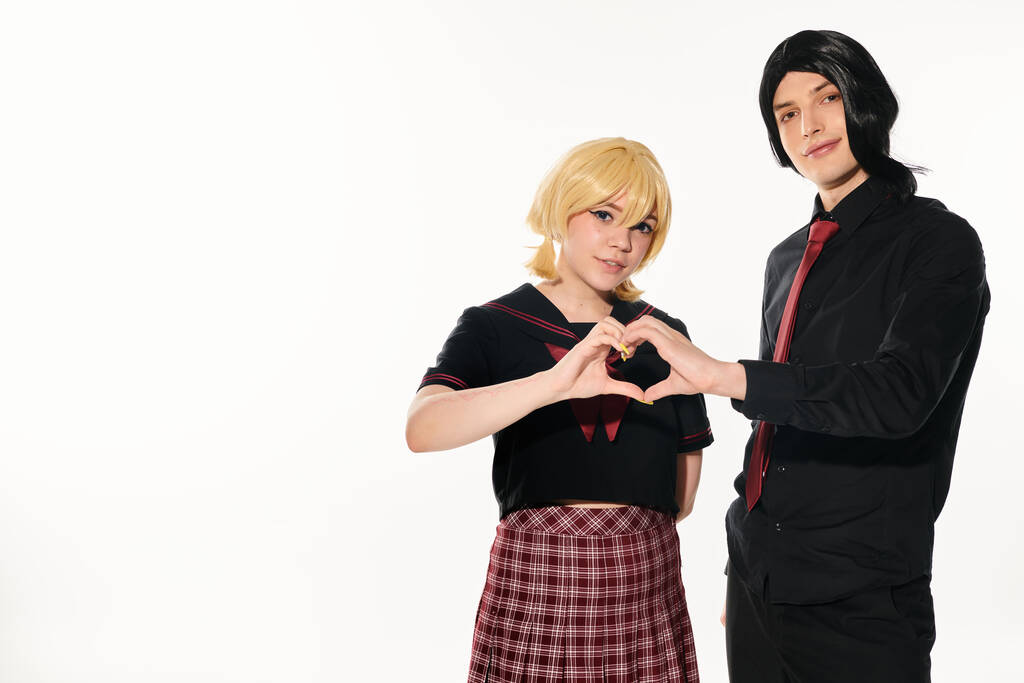 cosplay para w czarny i blond peruki pokazując znak serca z rąk patrząc na aparat fotograficzny na biały - Zdjęcie, obraz