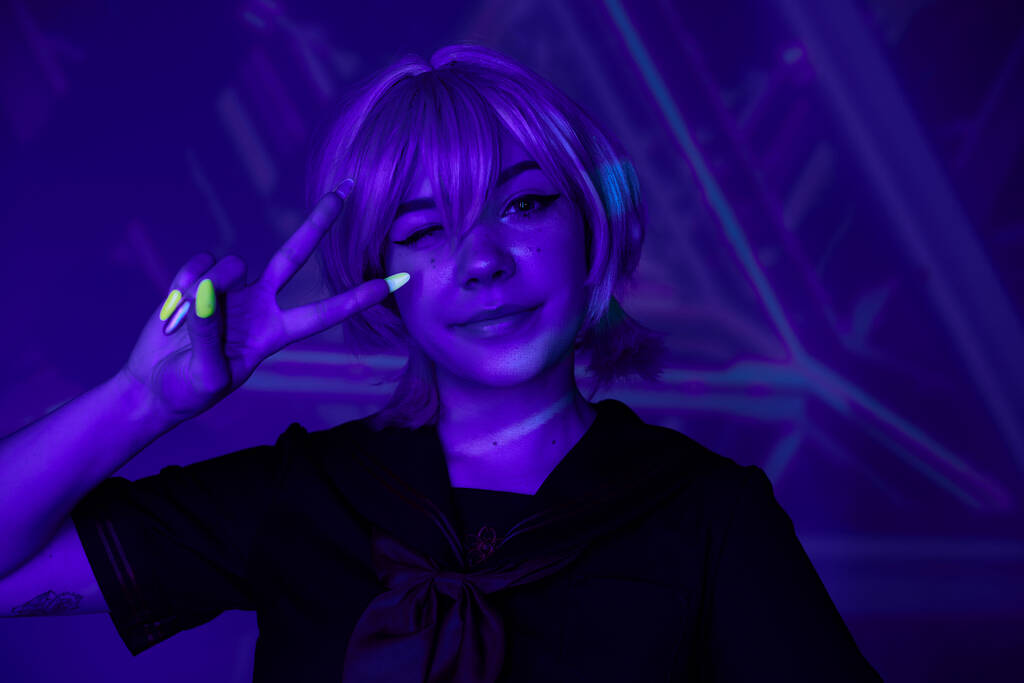 Blonde Anime-Frau mit fluoreszierender Maniküre zwinkert und zeigt Siegeszeichen in blauem Neonlicht - Foto, Bild