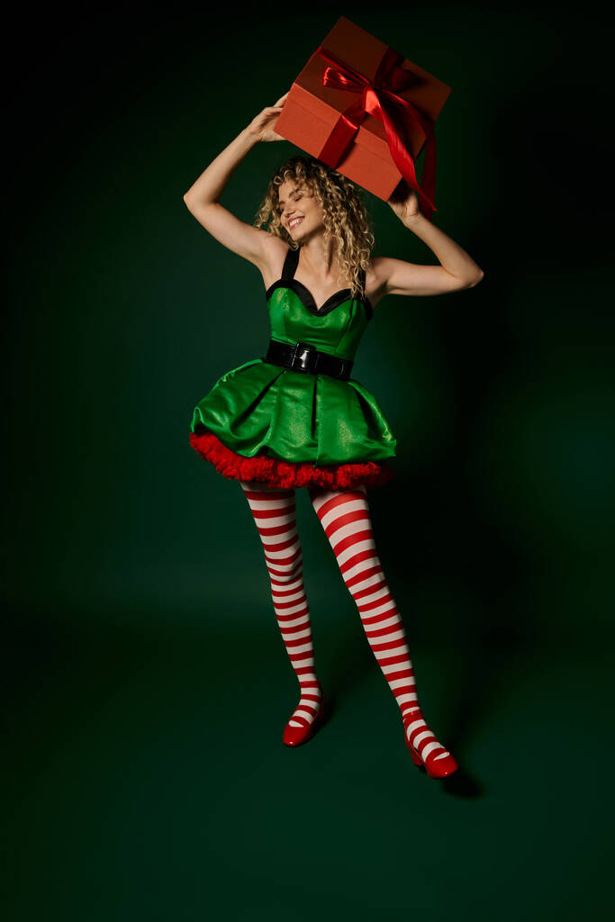 sonriente elfo de año nuevo en vestido verde festivo sosteniendo presente sobre su cabeza y mirando hacia otro lado - Foto, Imagen