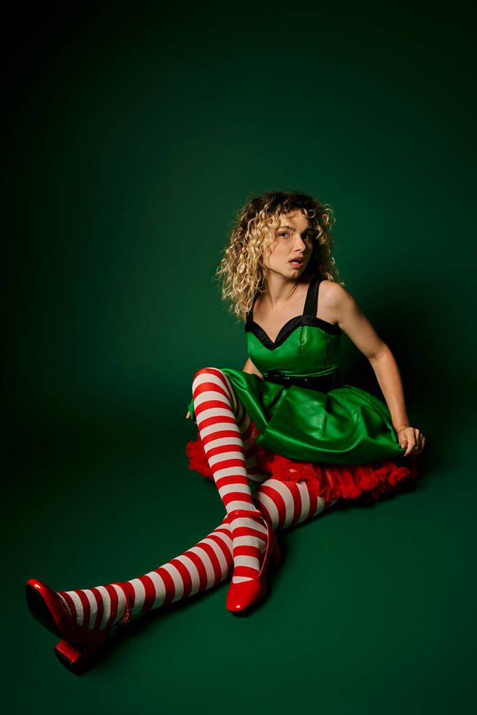 zasněný kudrnatý žena oblečená jako nový rok elf sedí na podlaze pózování na tmavě zeleném pozadí - Fotografie, Obrázek