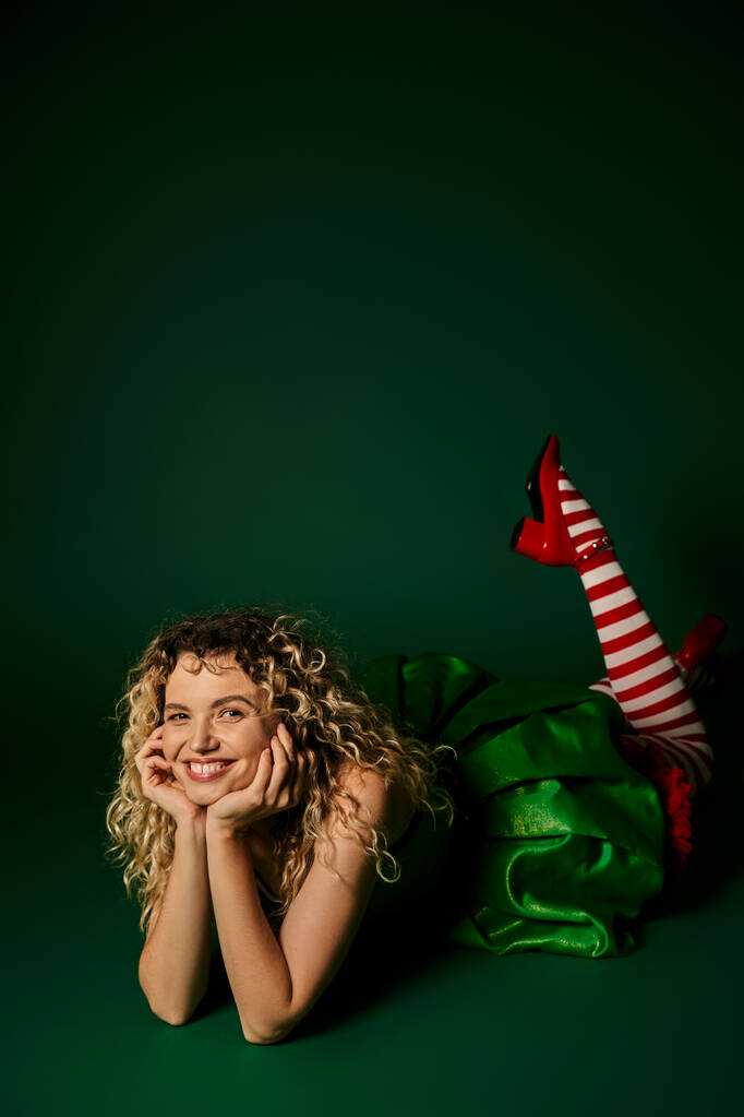 glückliche Frau, die als Neujahrselfe gekleidet auf dem Boden posiert, die Beine leicht erhöht auf grünem Hintergrund - Foto, Bild