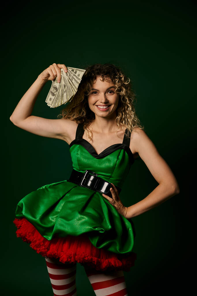 heureuse femme bouclée habillée comme elfe de nouvelle année avec un bras akimbo et avec de l'argent dans une autre main - Photo, image