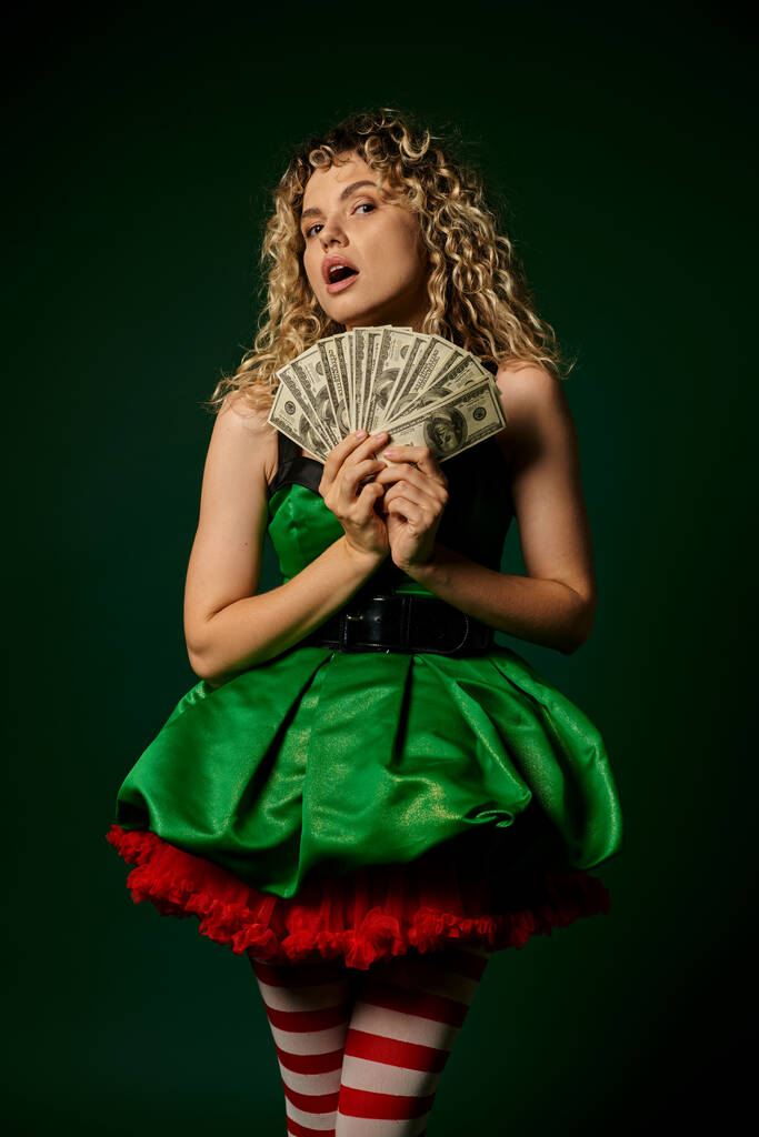 astonished pretty new year elf in green dress and striped stockings holding cash looking at camera - Photo, Image