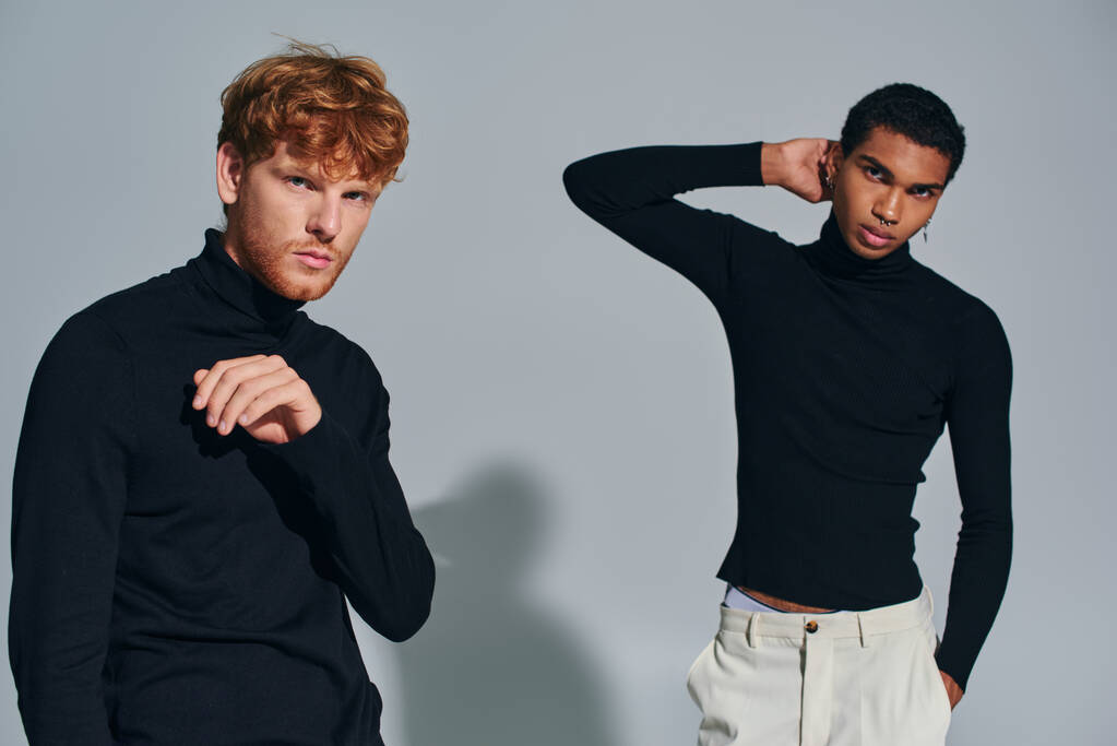 zwei multirassische männliche Modelle mit Zubehör in schwarzen Rollkragenpullovern gestikulierend und eine Kamera suchend - Foto, Bild