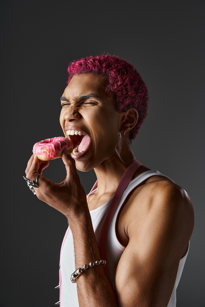voguish gutaussehender Mann mit rosa Haaren und Accessoires essen köstlichen rosa Donut, Mode und Stil - Foto, Bild