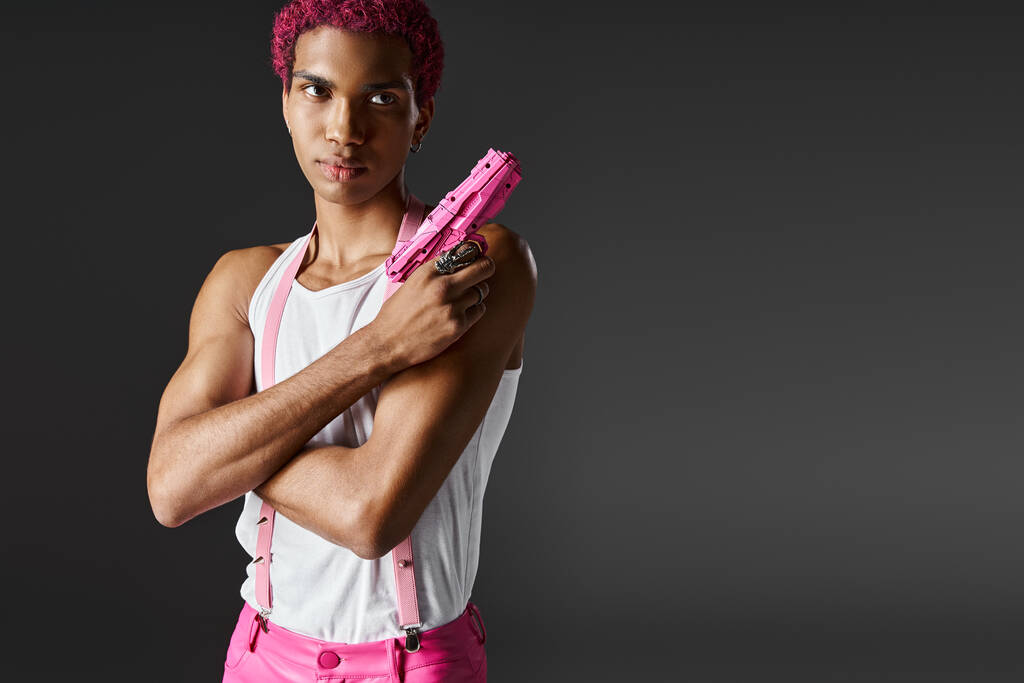 modelo masculino guapo de moda con pelo rosa posando con pistola de juguete mirando seriamente a la cámara - Foto, imagen