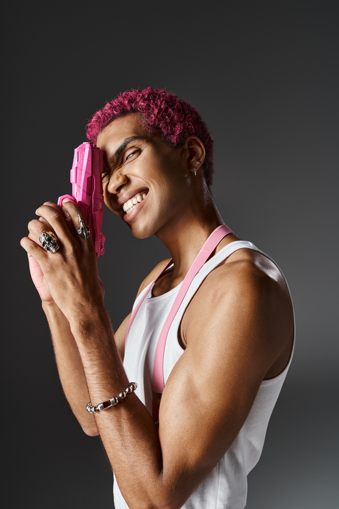 wesoły mężczyzna model z kręconymi różowymi włosami pozowanie w profilu z różowym pistoletem zabawki i uśmiech do kamery - Zdjęcie, obraz