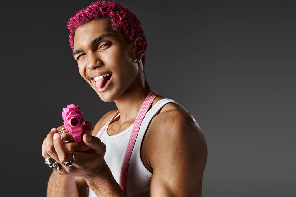 juguetona modelo masculino de pelo rosa sobresaliendo de su lengua y nivelando su pistola de juguete rosa en la cámara - Foto, imagen