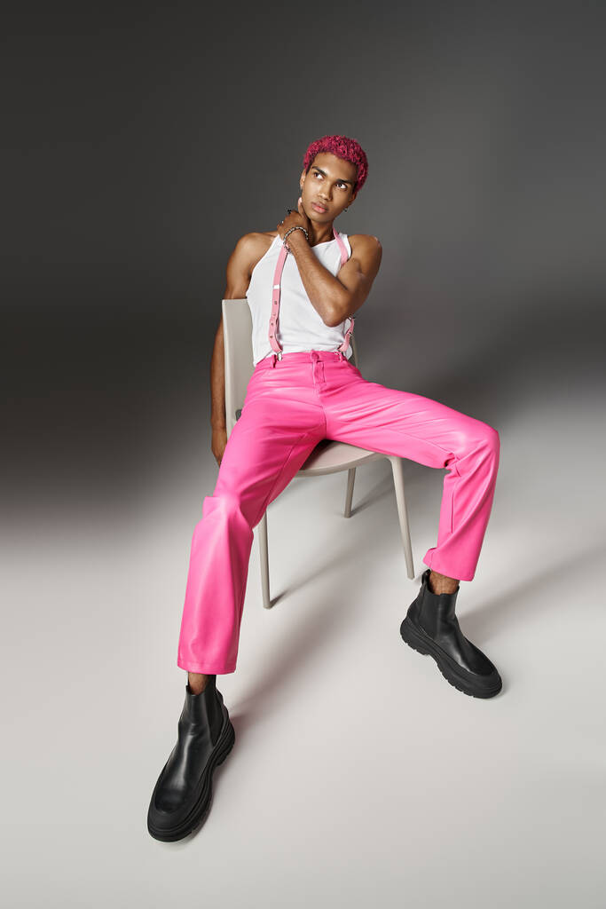 africký americký mužský model v růžových kalhotách s podvazky pózujícími na bílém křesle, módní koncept - Fotografie, Obrázek