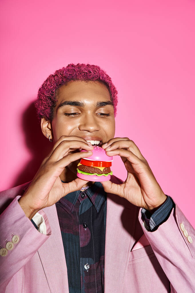boa aparência elegante modelo masculino com cabelo encaracolado comendo delicioso mini hambúrguer em pano de fundo rosa - Foto, Imagem