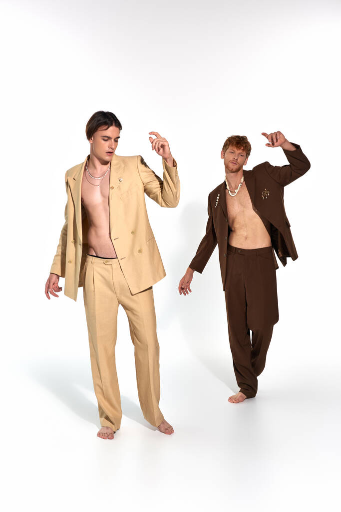 vertikale Aufnahme eines gut aussehenden eleganten Duos in aufgeknöpften Anzügen, das barfuß in Bewegungen posiert, Männer-Power - Foto, Bild