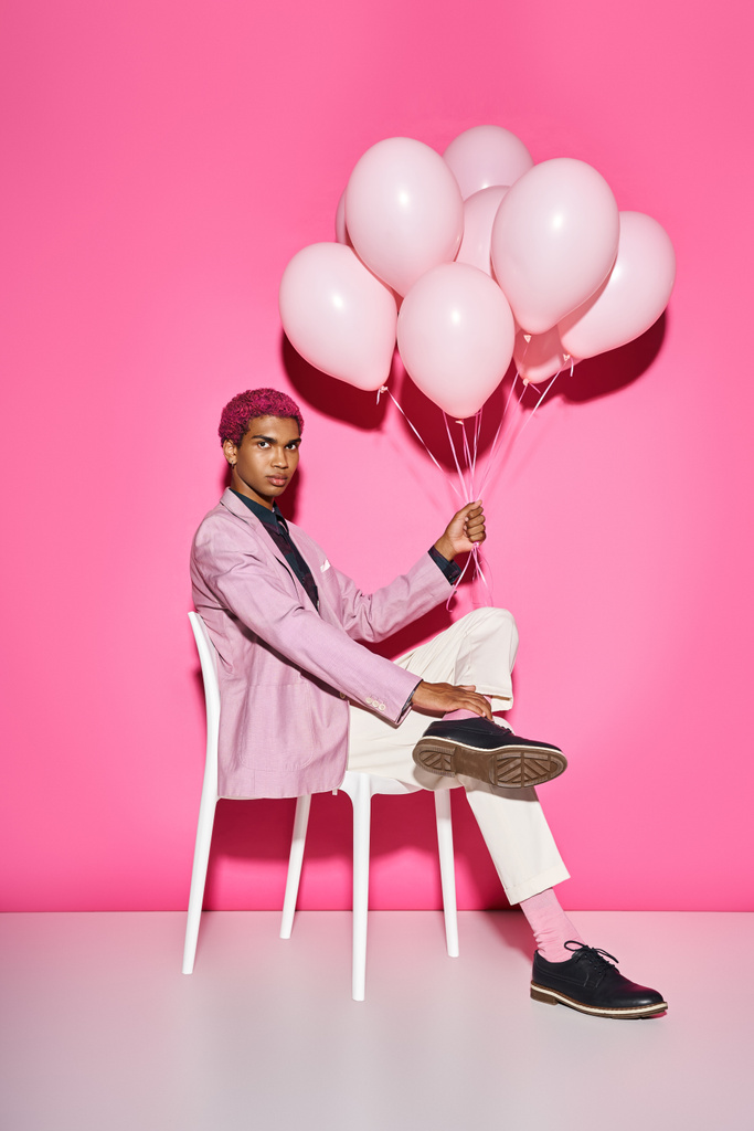εμφανίσιμος ροζ μαλλιά άνθρωπος κάθεται σε λευκή καρέκλα και κρατώντας μπαλόνια κοιτάζοντας κάμερα - Φωτογραφία, εικόνα