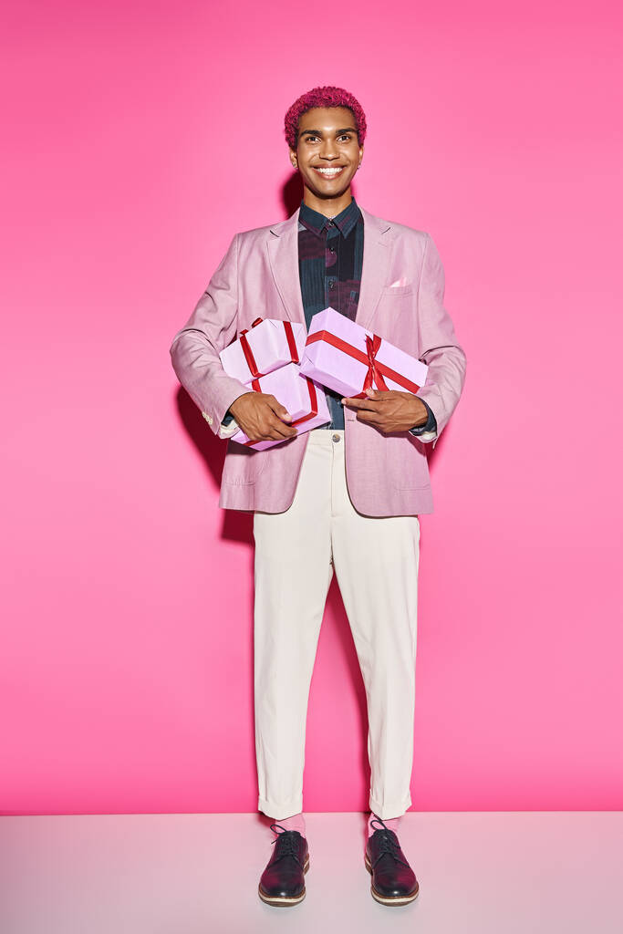fröhlicher junger Mann posiert unnatürlich mit Geschenken in den Händen auf rosa Hintergrund - Foto, Bild